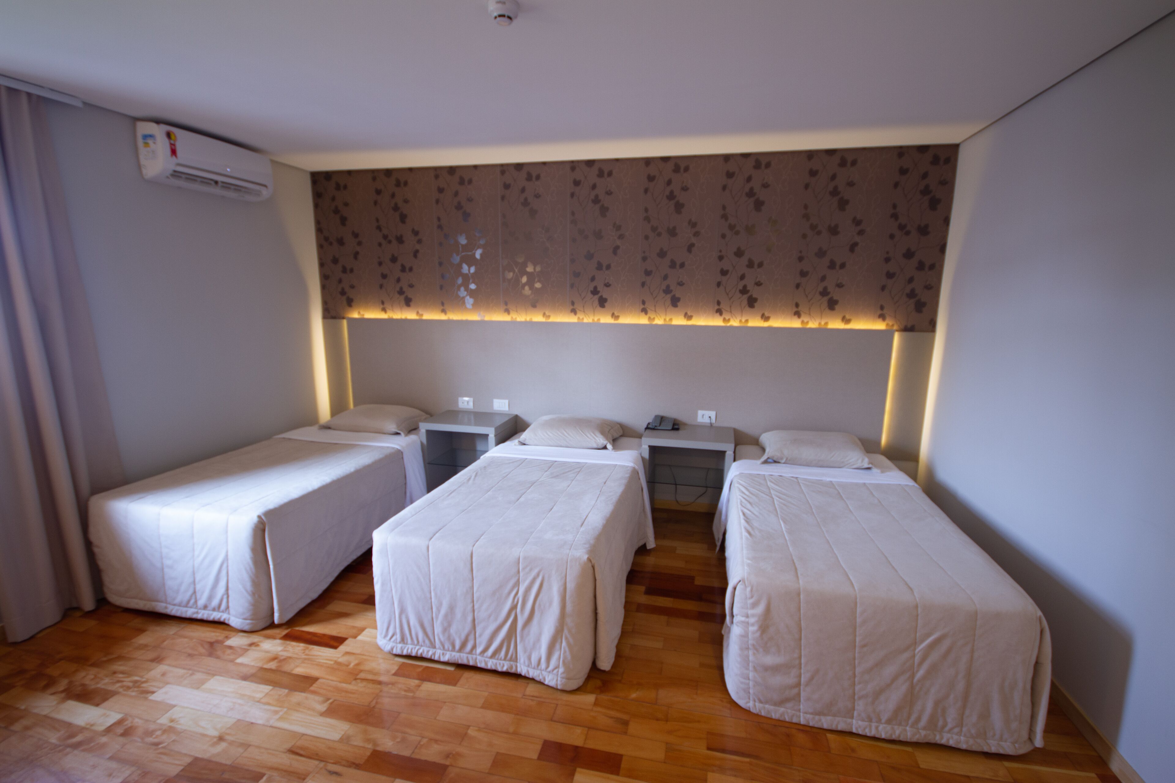 A hotel room with three single beds, a wooden floor, a patterned wall, and an air conditioning unit.