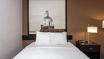 Ein Hotelzimmer mit einem Bett, einem Nachttisch mit Lampe und einem gerahmten Bild eines Leuchtturms an der Wand.