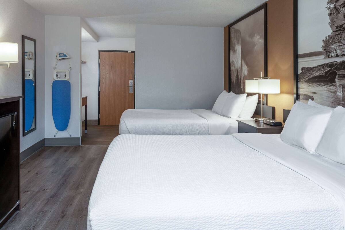 Une chambre d’hôtel avec deux lits, un bureau, une lampe, un miroir et une porte donnant sur une autre pièce.