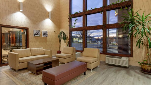 Un salon moderne avec un canapé beige, un pouf marron et une table basse. De grandes fenêtres équipées de stores laissent entrer la lumière, une plante en pot décore l’espace et une vue sur l’extérieur est offerte.