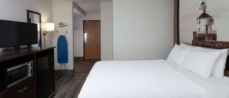 A hotel room with a large bed, a flat-screen TV, a dresser, a microwave, and a painting of a lighthouse on the wall.