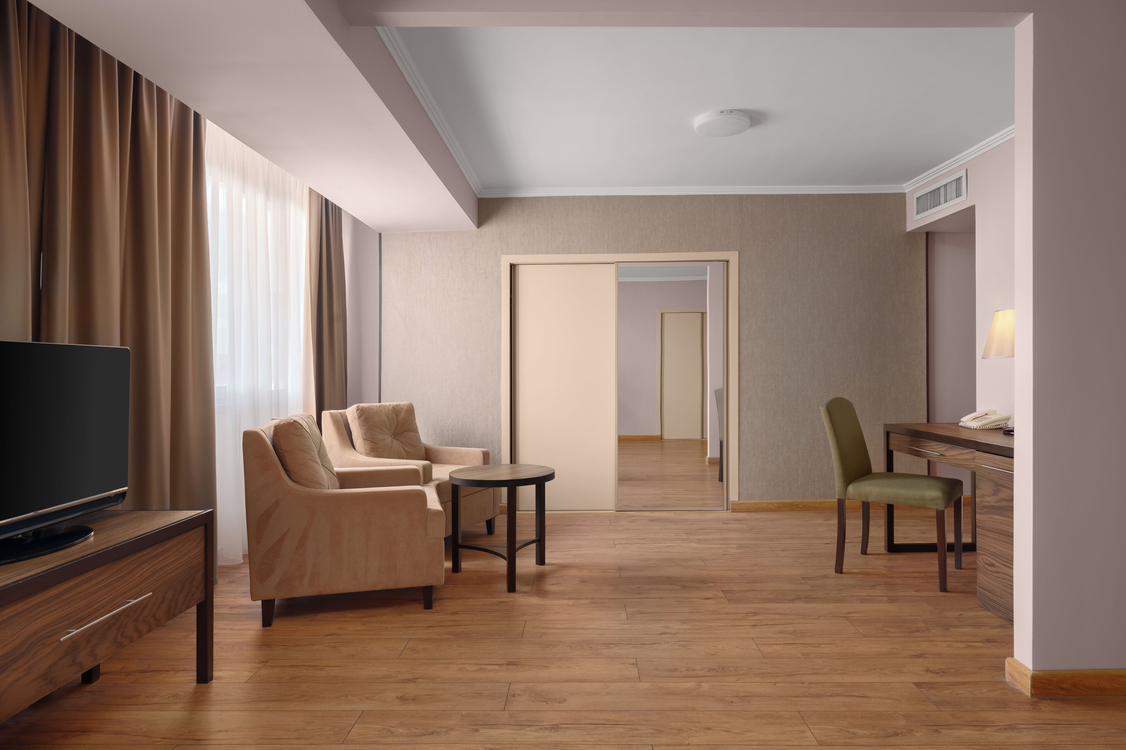 A hotel room with a TV, sofa, armchair, small table, desk, chair, and a mirror.