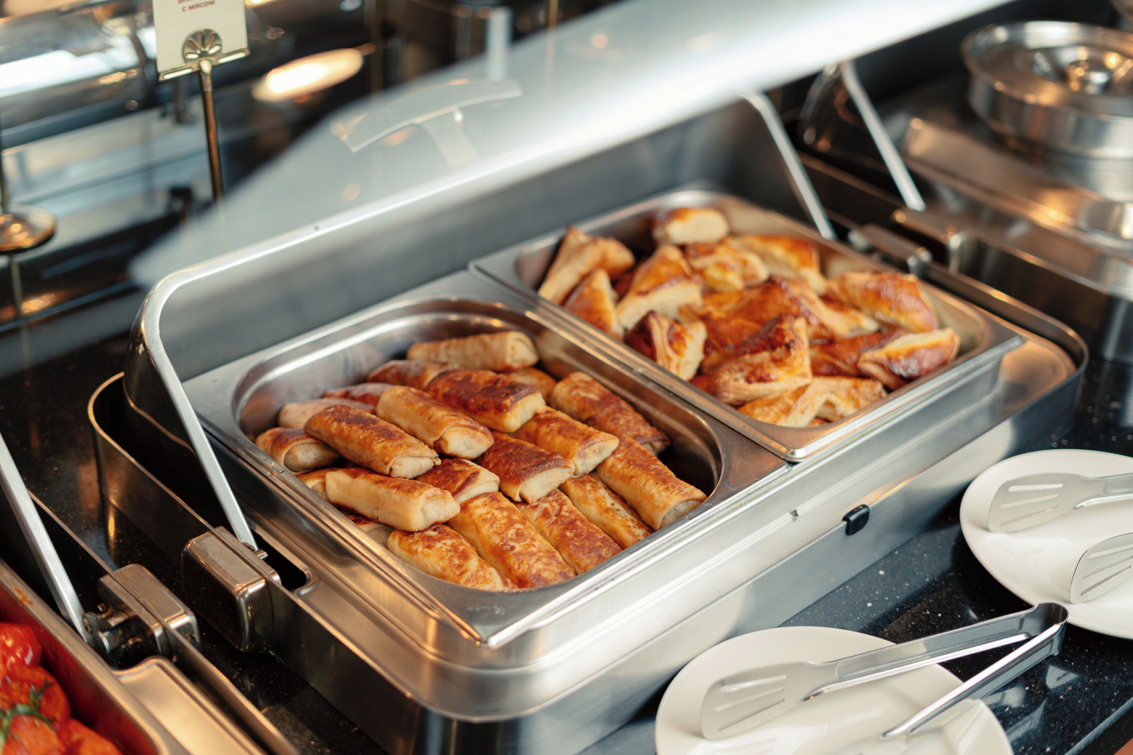A buffet with trays of food, serving utensils, and a stainless steel container.
