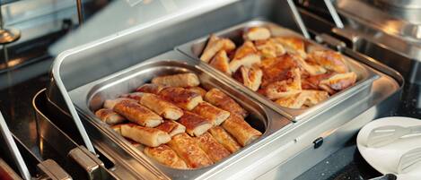 A buffet with trays of food, serving utensils, and a stainless steel container.