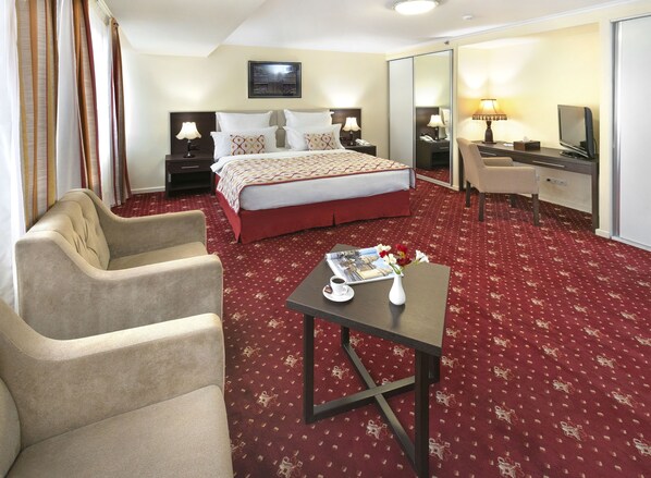 Habitación de hotel con una cama grande, dos mesitas de noche con lámparas, un escritorio con una silla, una pequeña mesa con un florero y una revista, y un espejo grande.