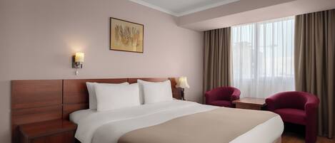 Habitación de hotel con una cama grande, dos sillones, una mesita, una mesita de noche, una lámpara de pared, un cuadro y una ventana con cortinas.