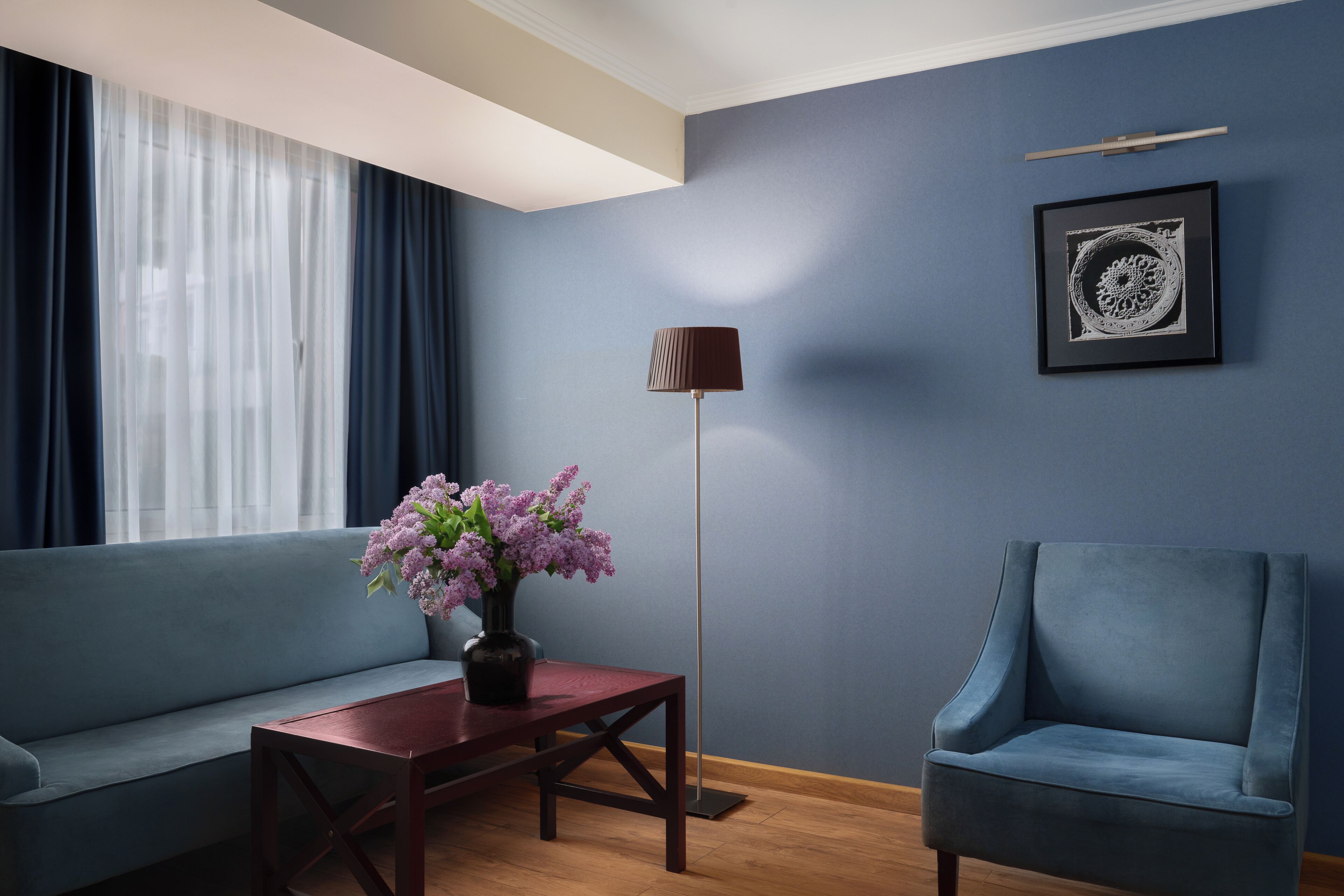 A room with a blue sofa, a wooden coffee table with a vase of flowers, a blue armchair, a floor lamp, and a framed picture on the wall.