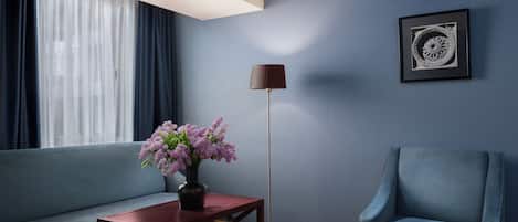 Una habitación con un sofá azul, una mesa de centro de madera con un jarrón de flores, un sillón azul, una lámpara de pie y un cuadro en la pared.