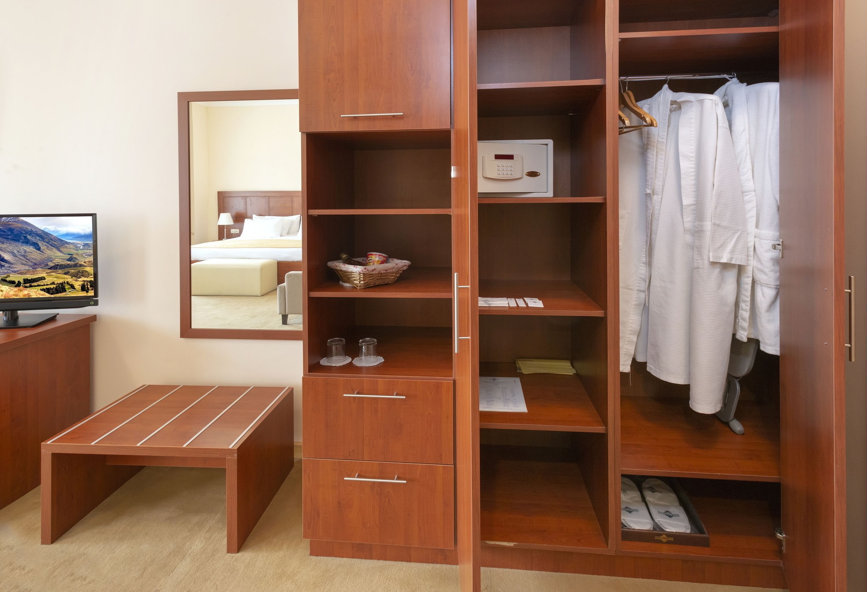 Habitación de hotel con un armario de madera, un pequeño banco de madera, televisión y un espejo que refleja la habitación.