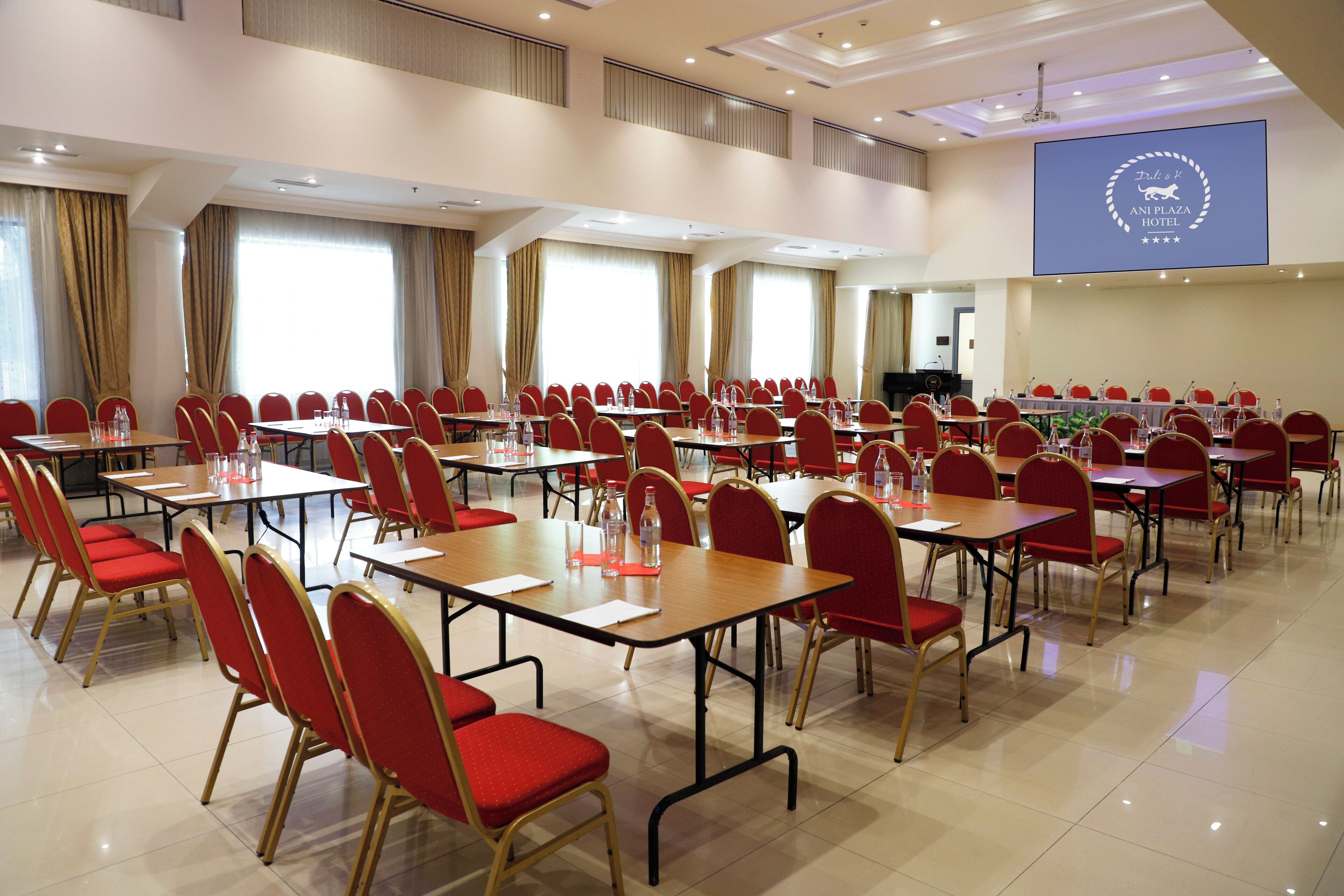 A conference room with multiple tables and red chairs, large windows, and a screen displaying the hotel name.