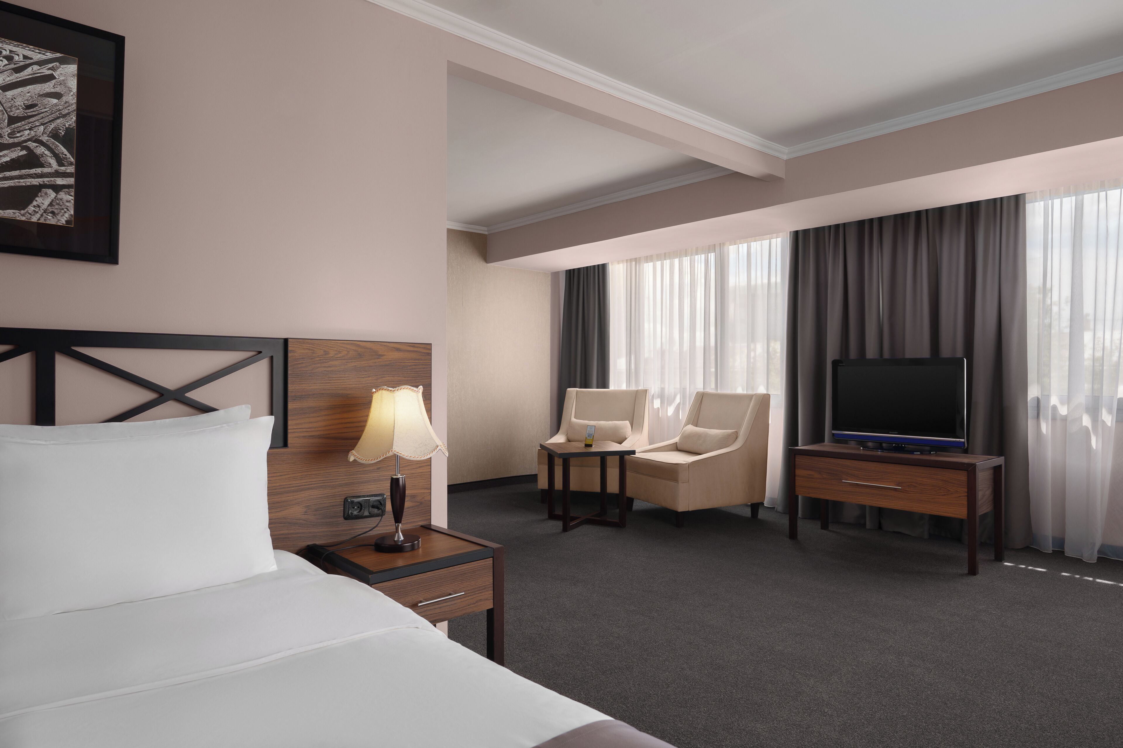 Habitación de hotel con una cama, un escritorio con lámpara, dos sillas, un televisor y un amplio ventanal con cortinas.