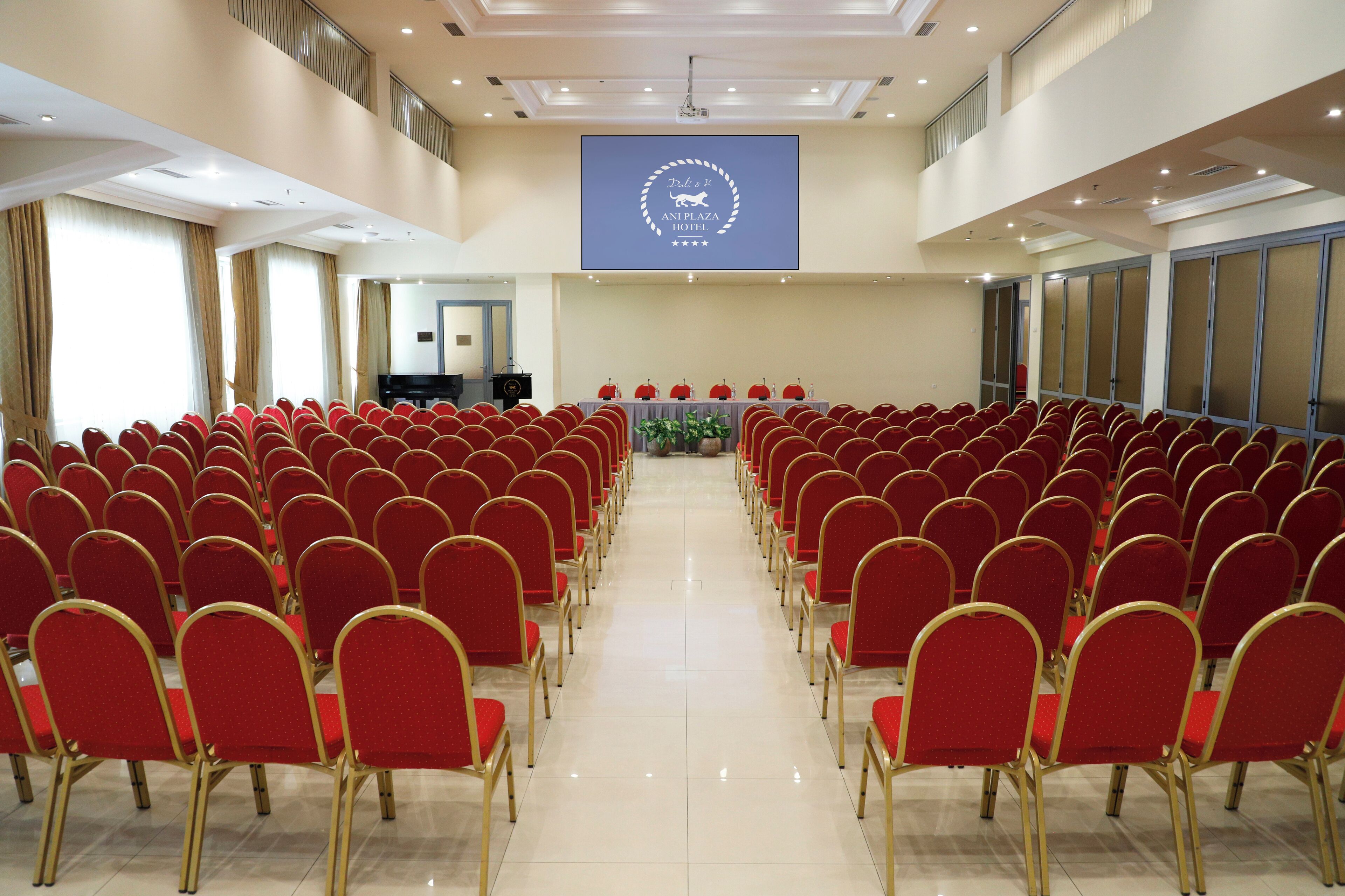 A conference room with rows of red chairs, a podium, and a large screen displaying the hotel name.