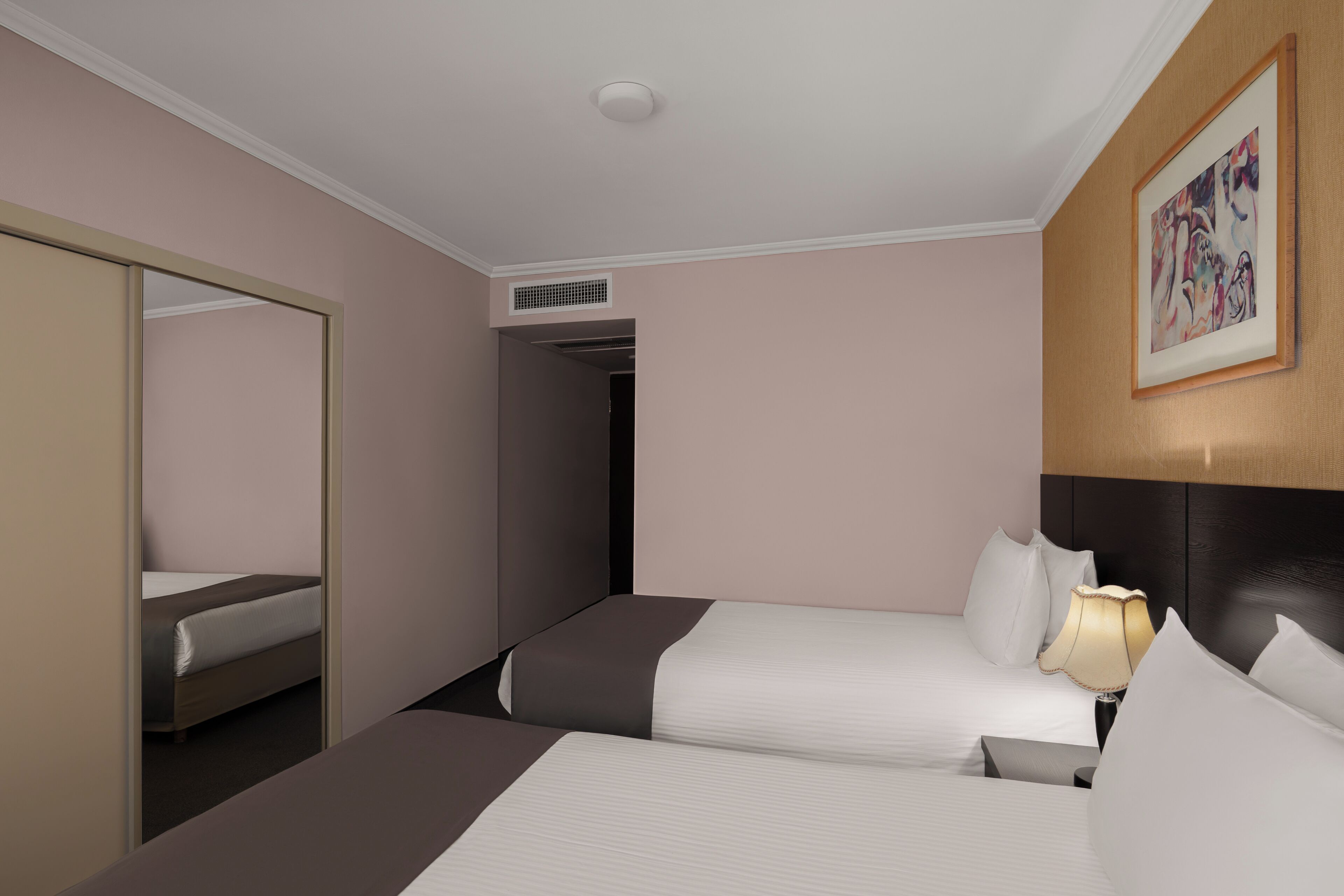 A hotel room with two beds, a large mirror, a painting on the wall, and a ceiling vent.
