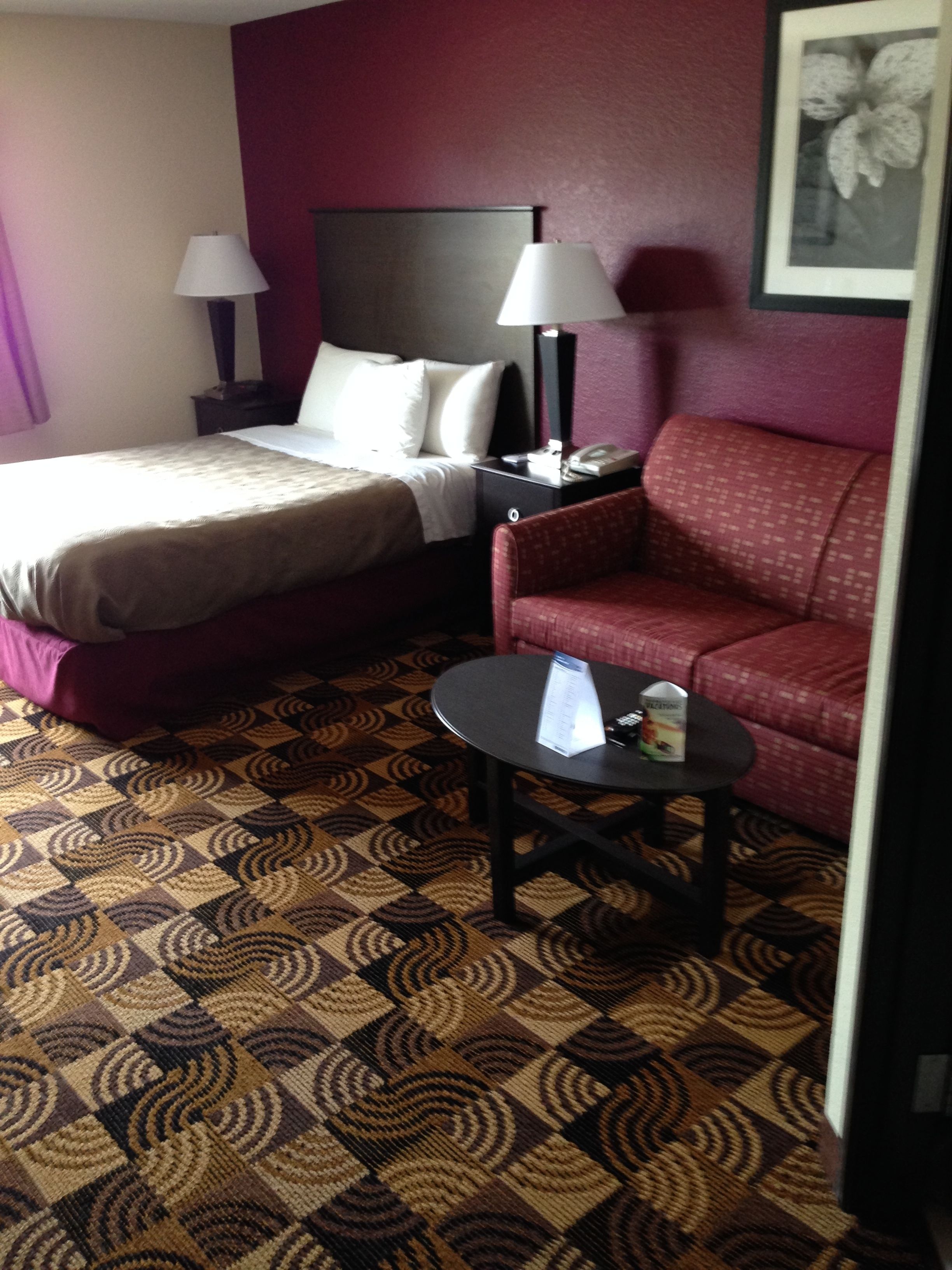 Una habitación de hotel con una cama, un sillón rojo, una mesita con un menú y una mesita de noche con una lámpara.