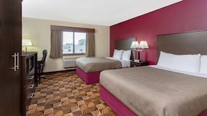 Habitación de hotel con dos camas, un escritorio, una silla, un ventanal con cortinas y una alfombra estampada.