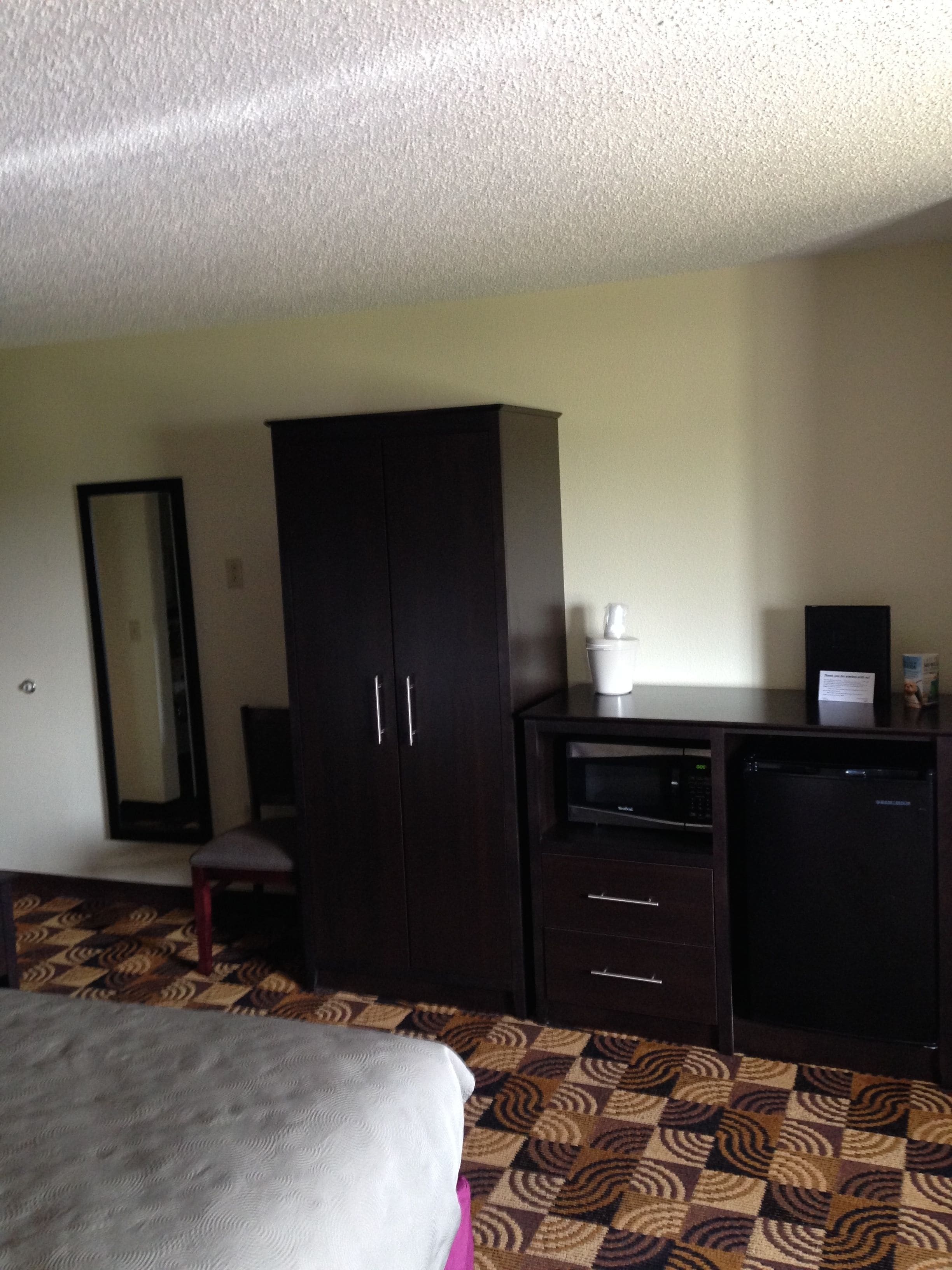 A hotel room with a bed, a dark wooden wardrobe, a small desk with a microwave, and a patterned carpet.