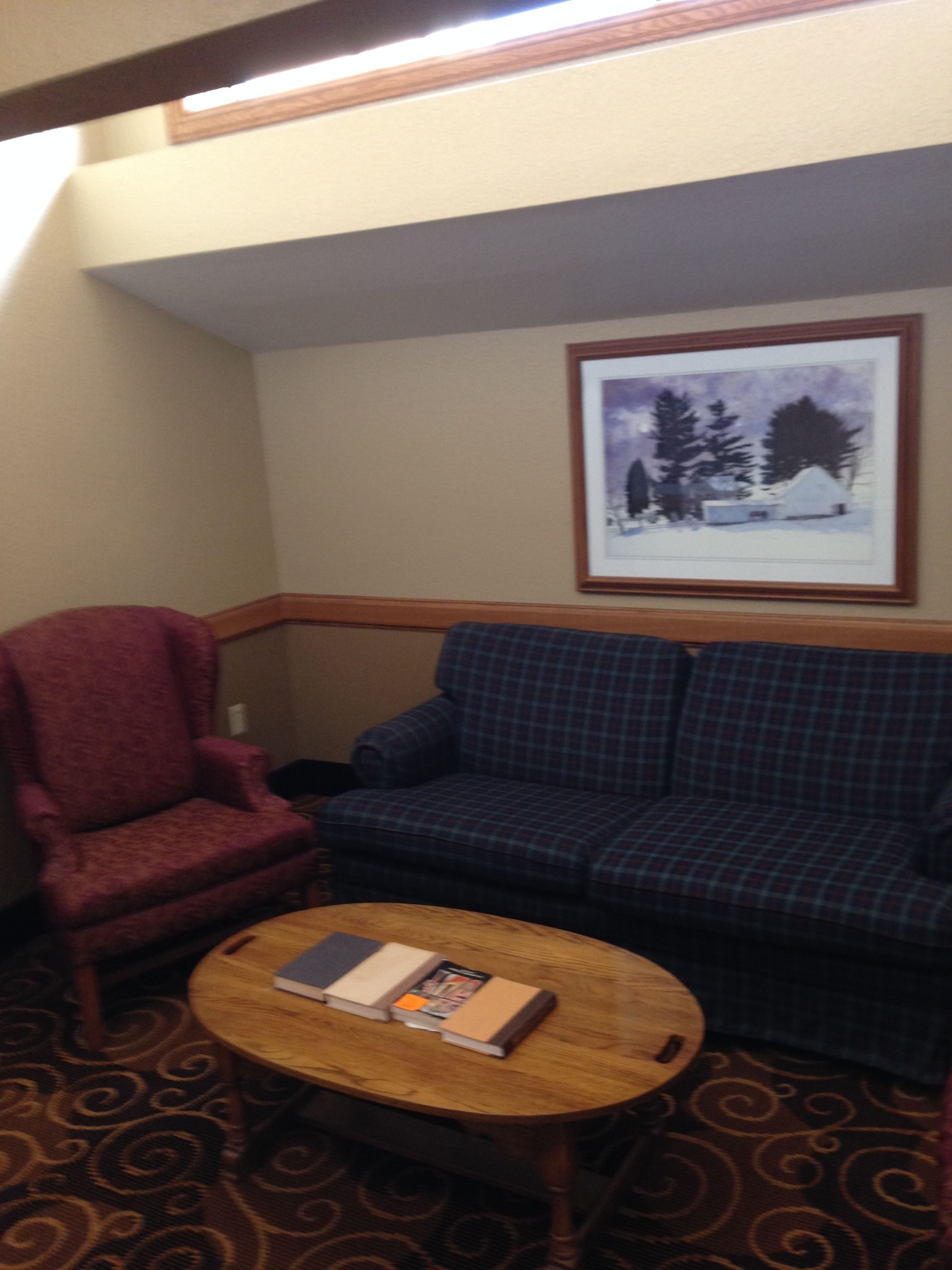 A room with a red armchair, a blue plaid sofa, and a wooden coffee table with books. A framed picture of a snowy landscape hangs on the wall.