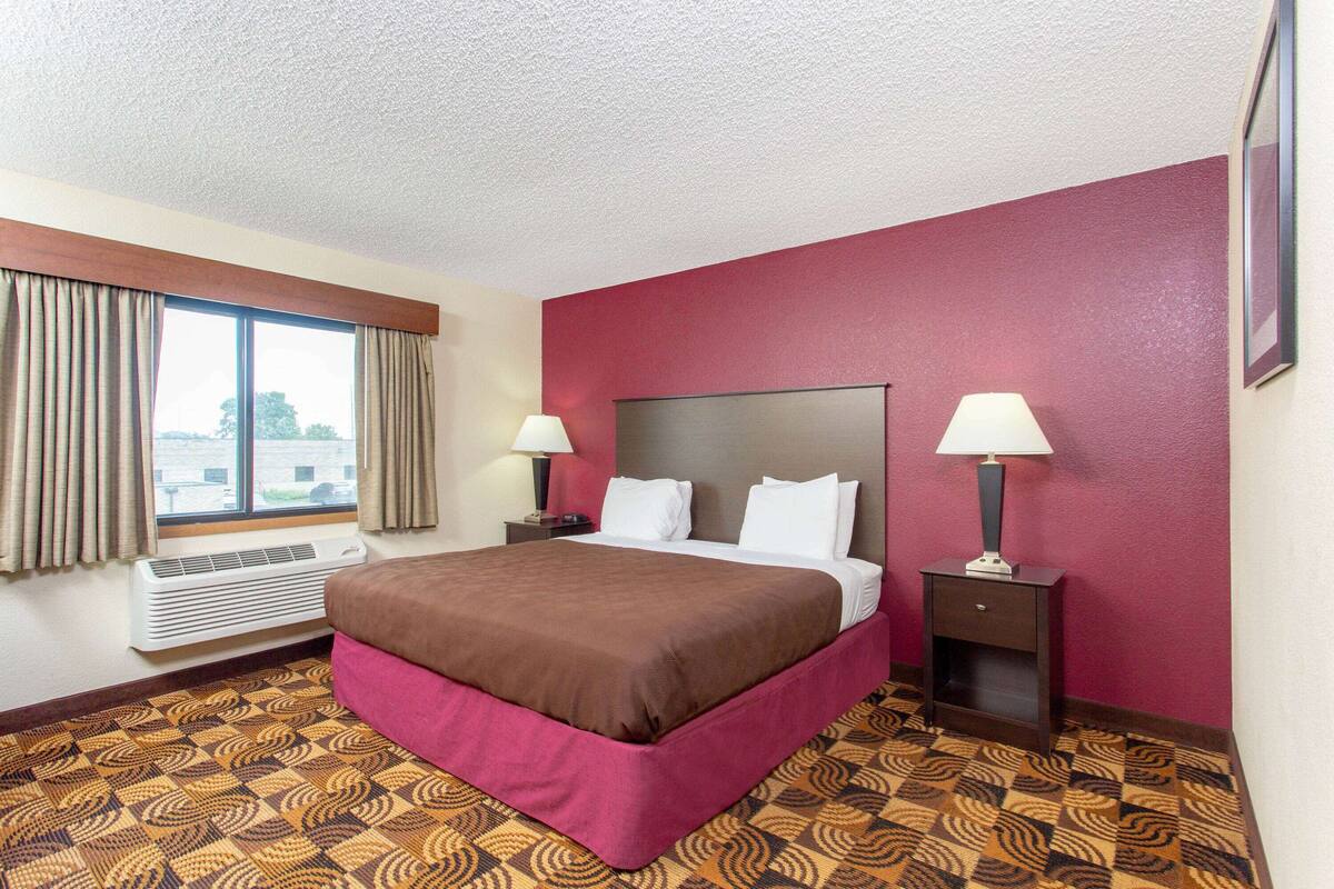 A hotel room with a large bed, two bedside tables with lamps, a window with curtains, and patterned carpet.