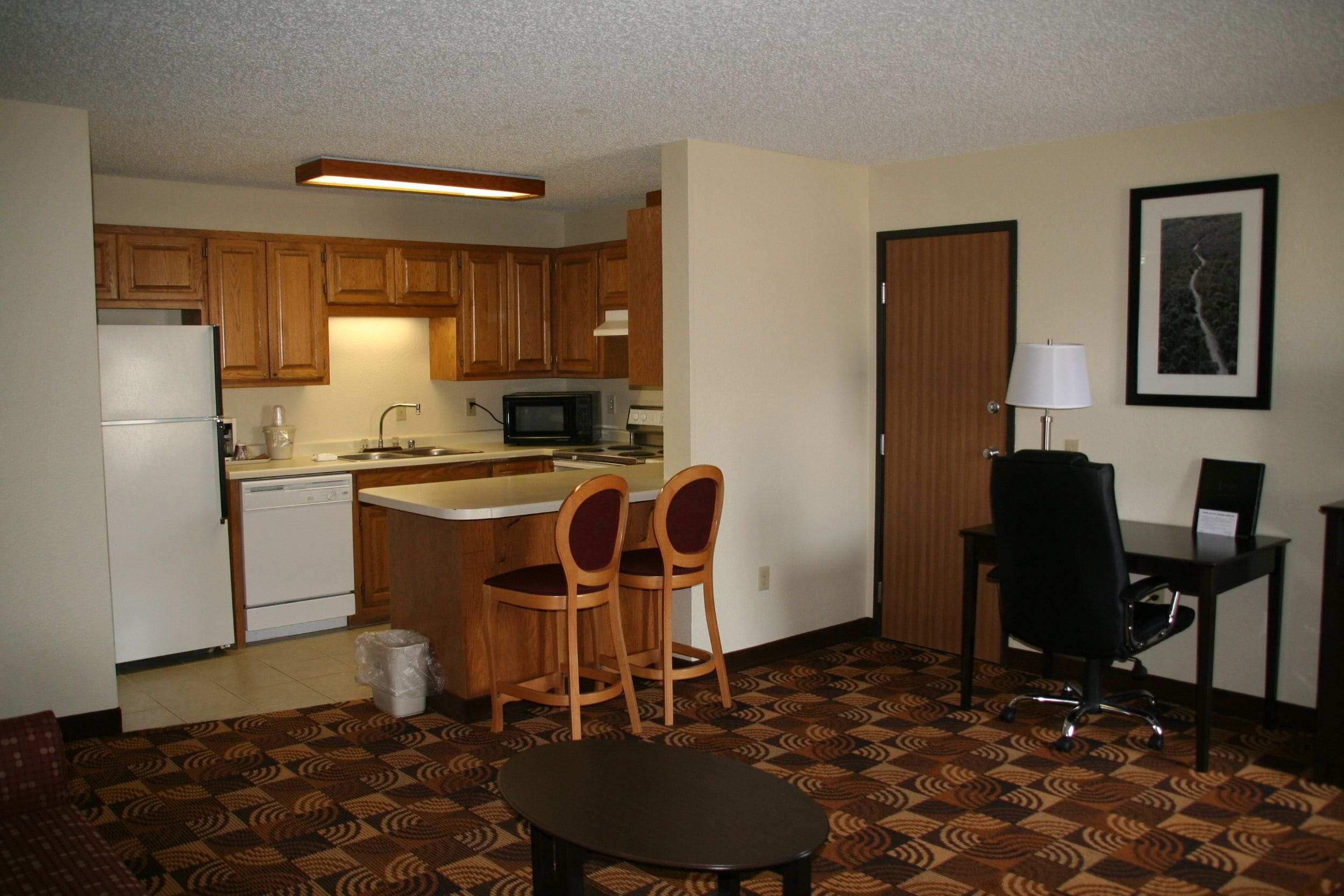 A compact kitchen with wooden cabinets, a microwave, and a refrigerator. There is a small dining area with two chairs and a table. The floor has a patterned carpet.