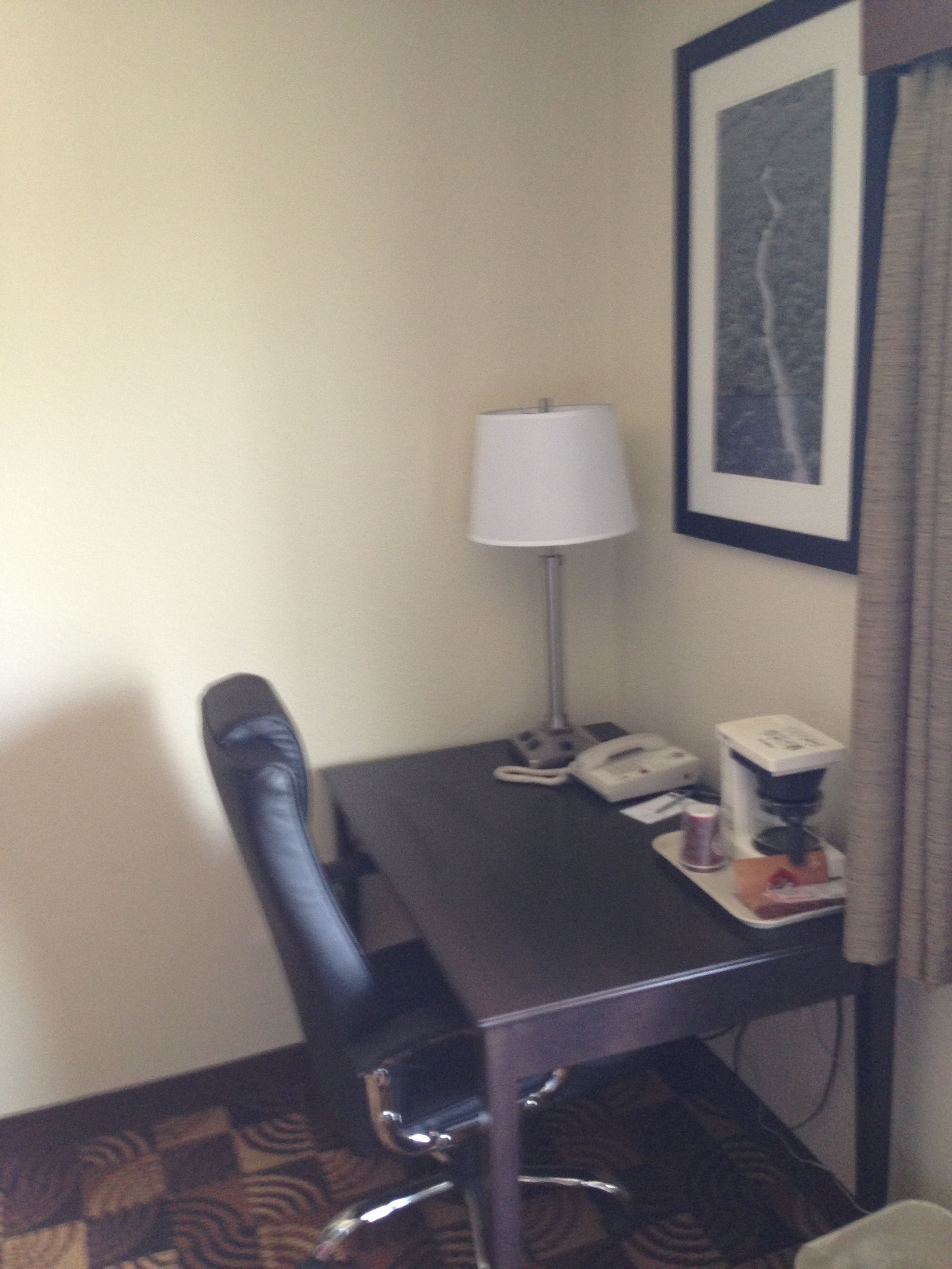 A hotel room with a desk, chair, lamp, phone, coffee maker, and a framed picture on the wall.