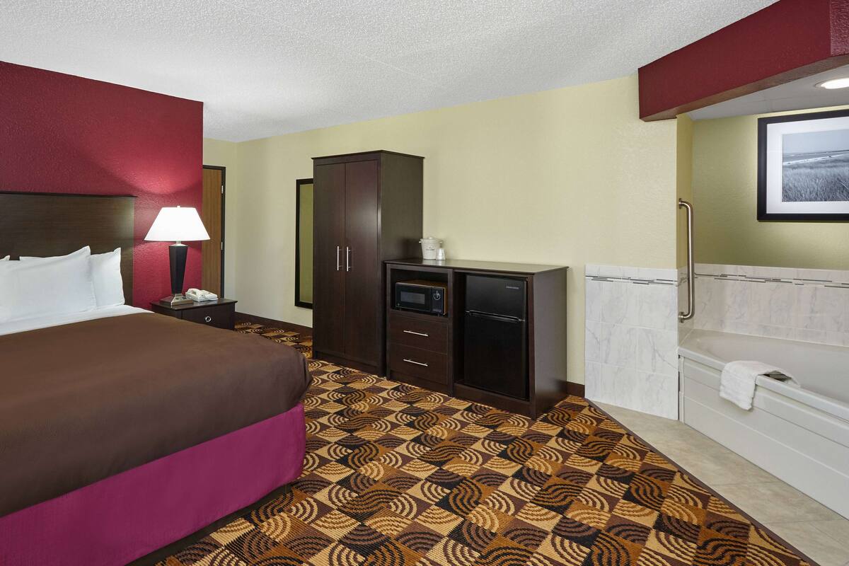 A hotel room with a bed, a desk with a lamp, a wardrobe, a microwave, and a bathroom with a bathtub.