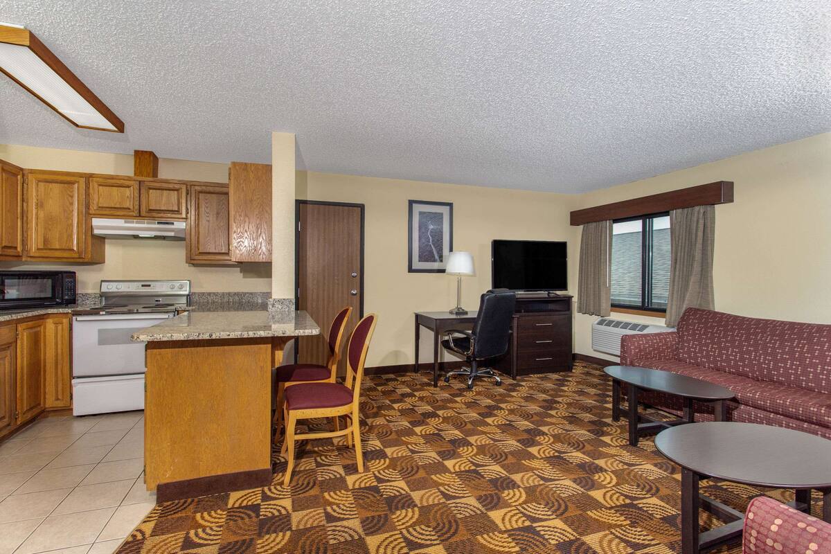 A compact hotel room with a kitchenette, a dining area, a sofa, and a TV.