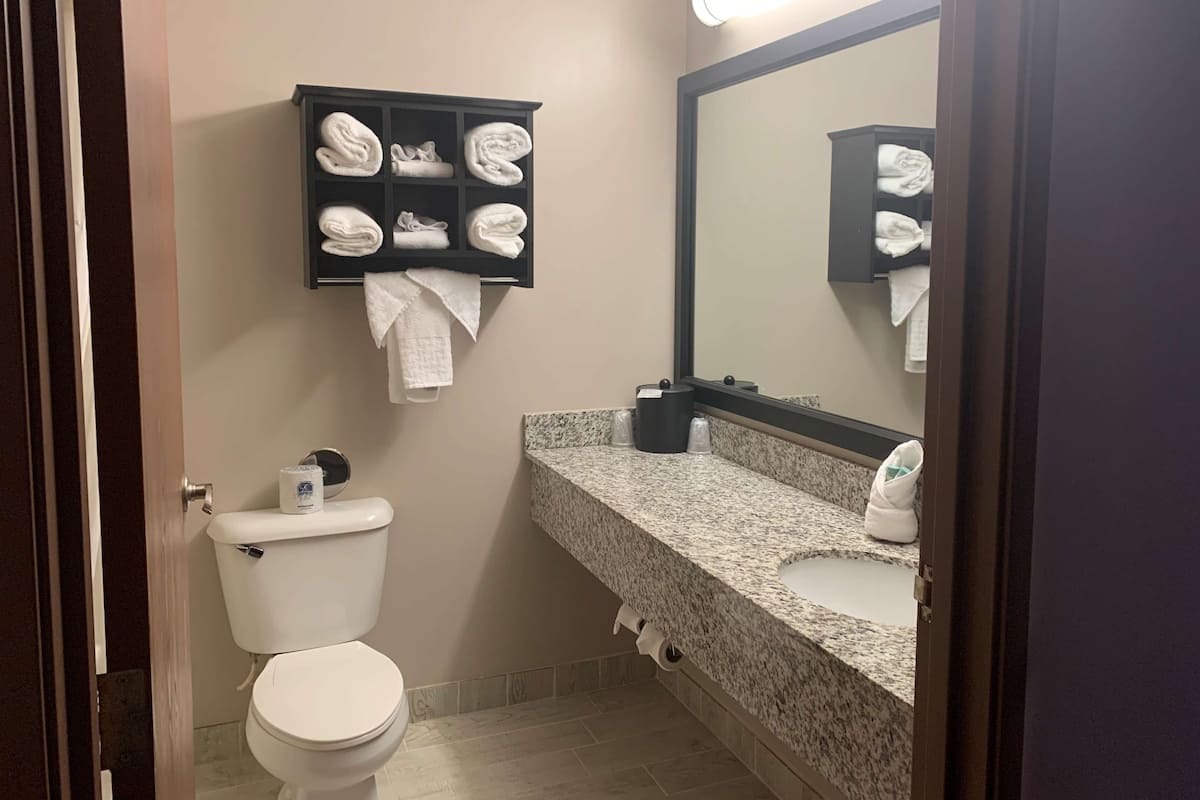 A bathroom with a toilet, a towel rack with folded towels, a sink with a mirror, and a trash can.