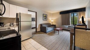 A compact hotel room with a kitchenette, a sofa, a small table, and a window with curtains.