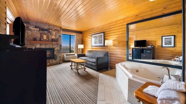 A log cabin interior with a stone fireplace, a sofa, a coffee table, a large bathtub, and a television.