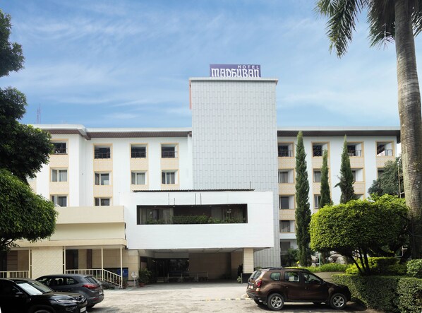 A hotel with a modern design, featuring a central tower with a sign, multiple balconies, and a parking area with cars. - Hotel Madhuban (Dehradun)