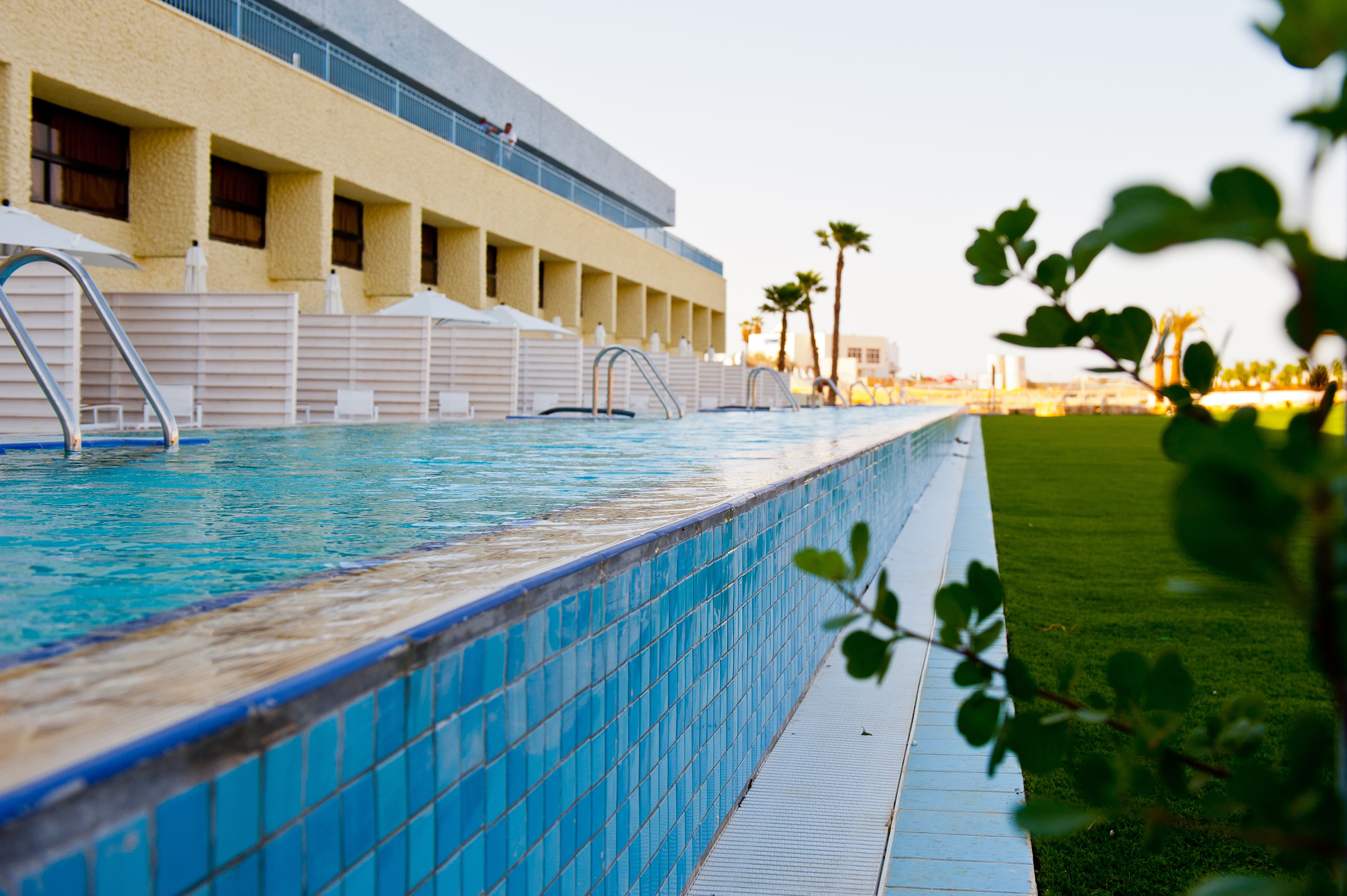 En swimmingpool med kakelbeklädd kant, en byggnad med balkonger och ett grönområde med palmer.