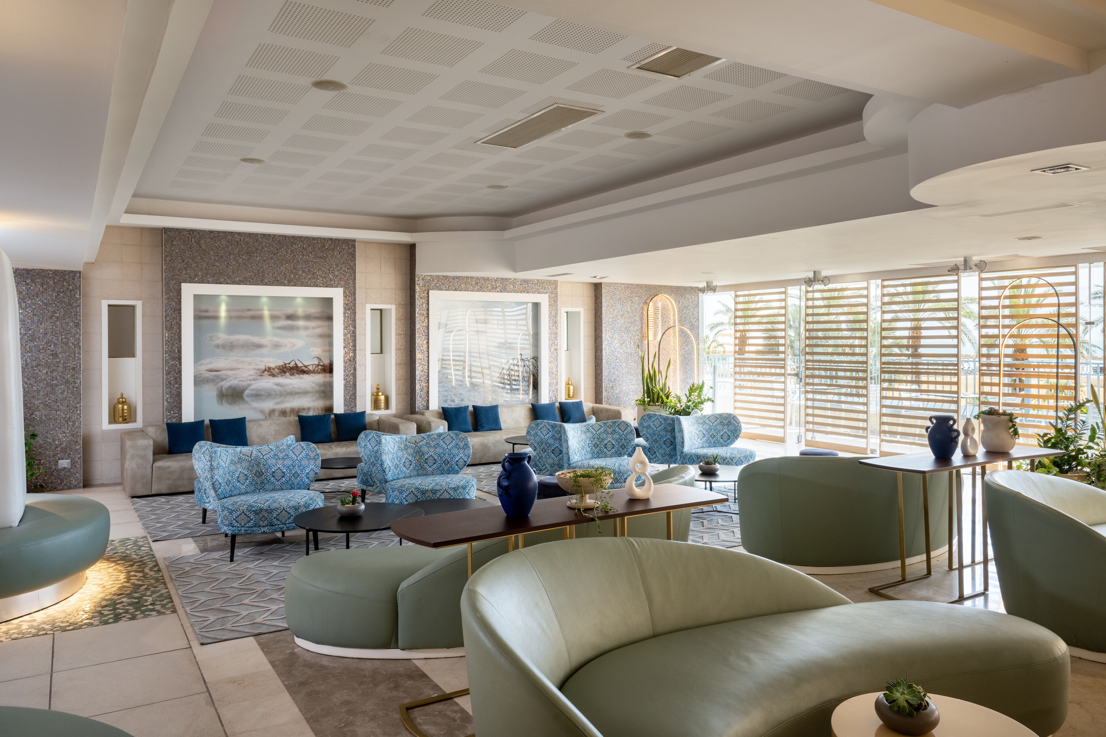 A modern hotel lobby with a seating area featuring blue patterned sofas and green armchairs, a coffee table, and a large window with blinds.