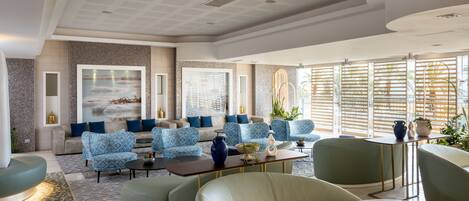 A modern hotel lobby with a seating area featuring blue patterned sofas and green armchairs, a coffee table, and a large window with blinds.