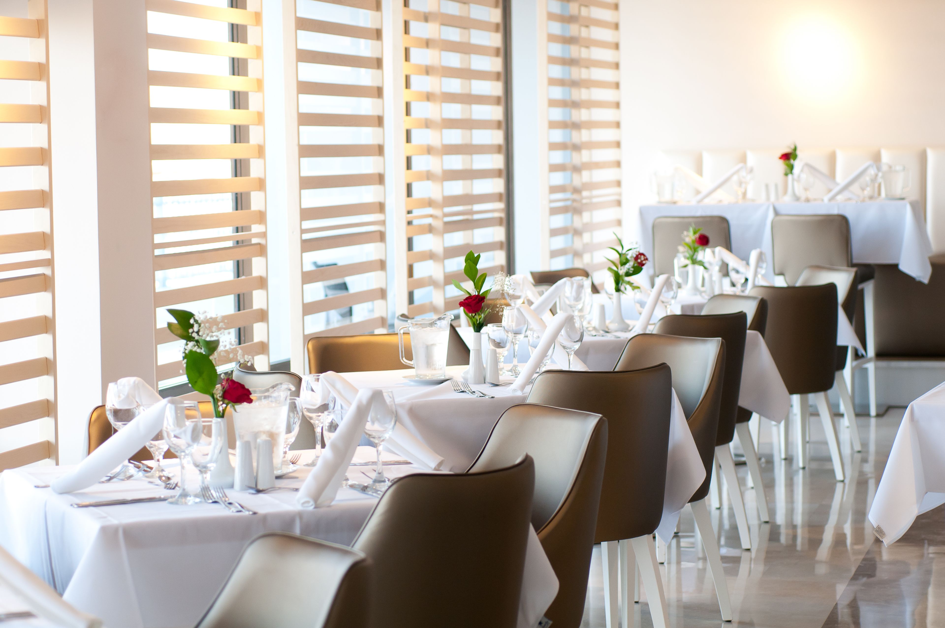 Un restaurant où les tables sont dressées pour le service, avec des nappes blanches, des chaises marron et des centres de table floraux.