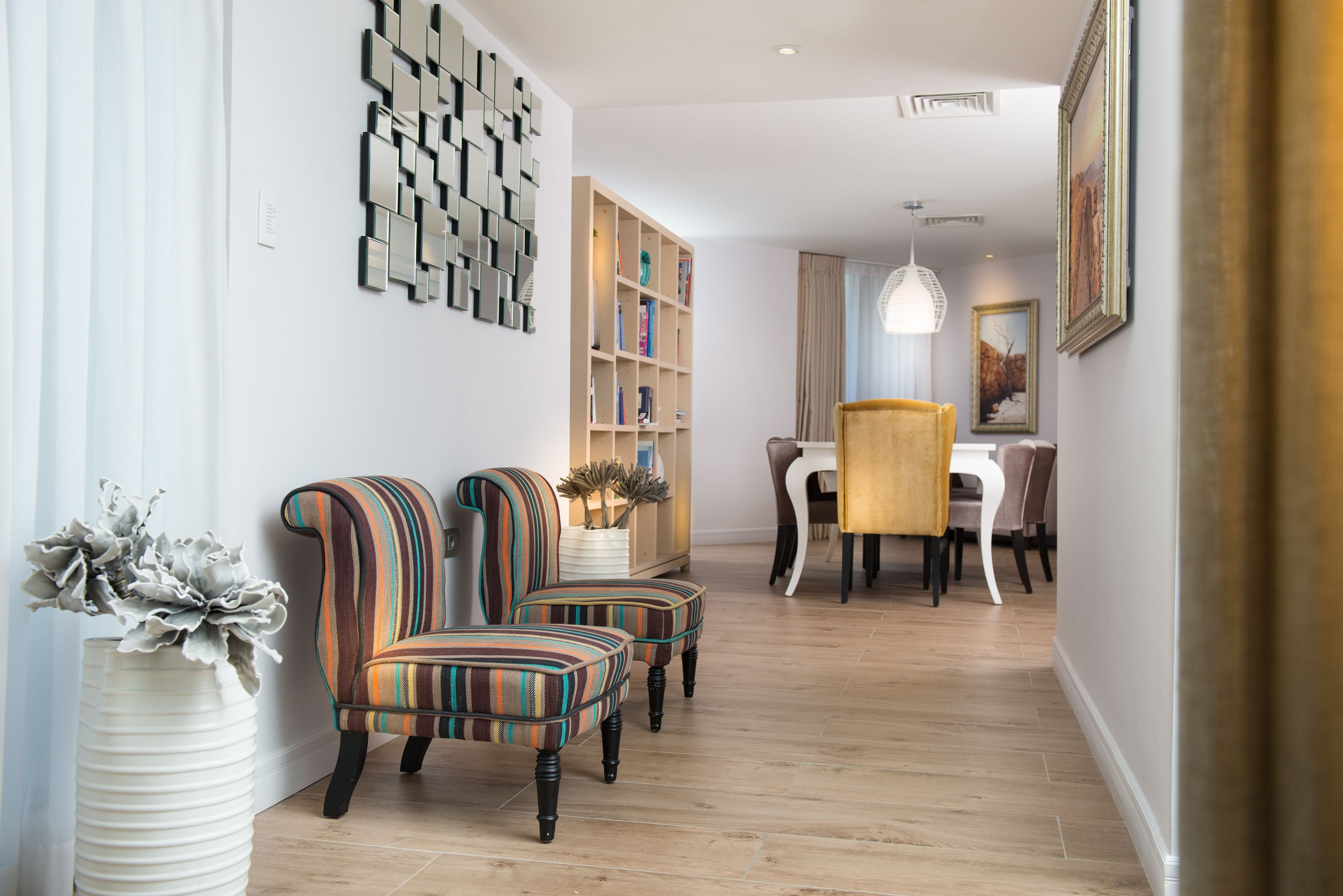 A modern living room with striped armchairs, a wooden floor, a bookshelf, and a dining area with a table and chairs.