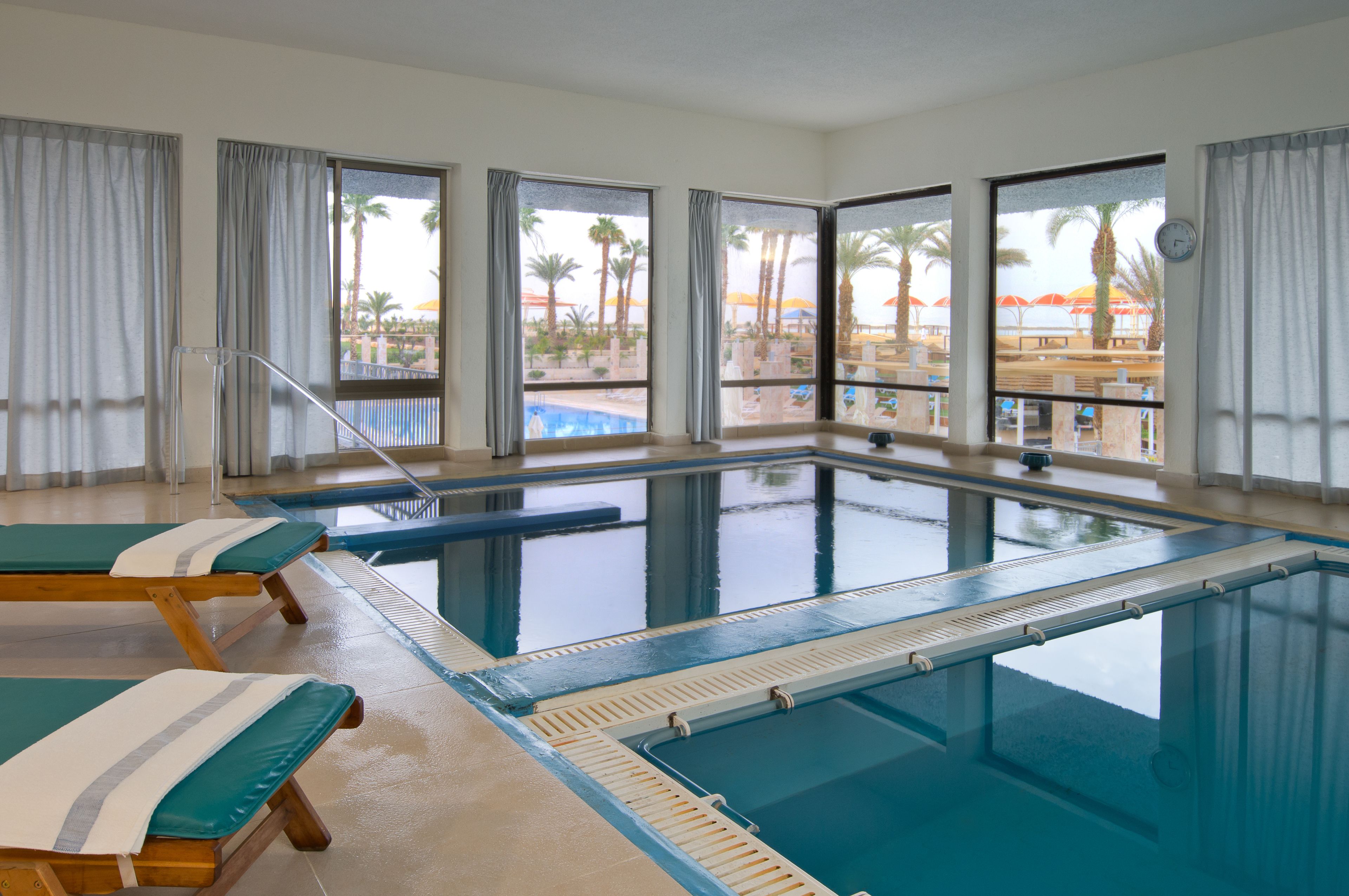 A swimming pool with clear water, two lounge chairs, and large windows with curtains.