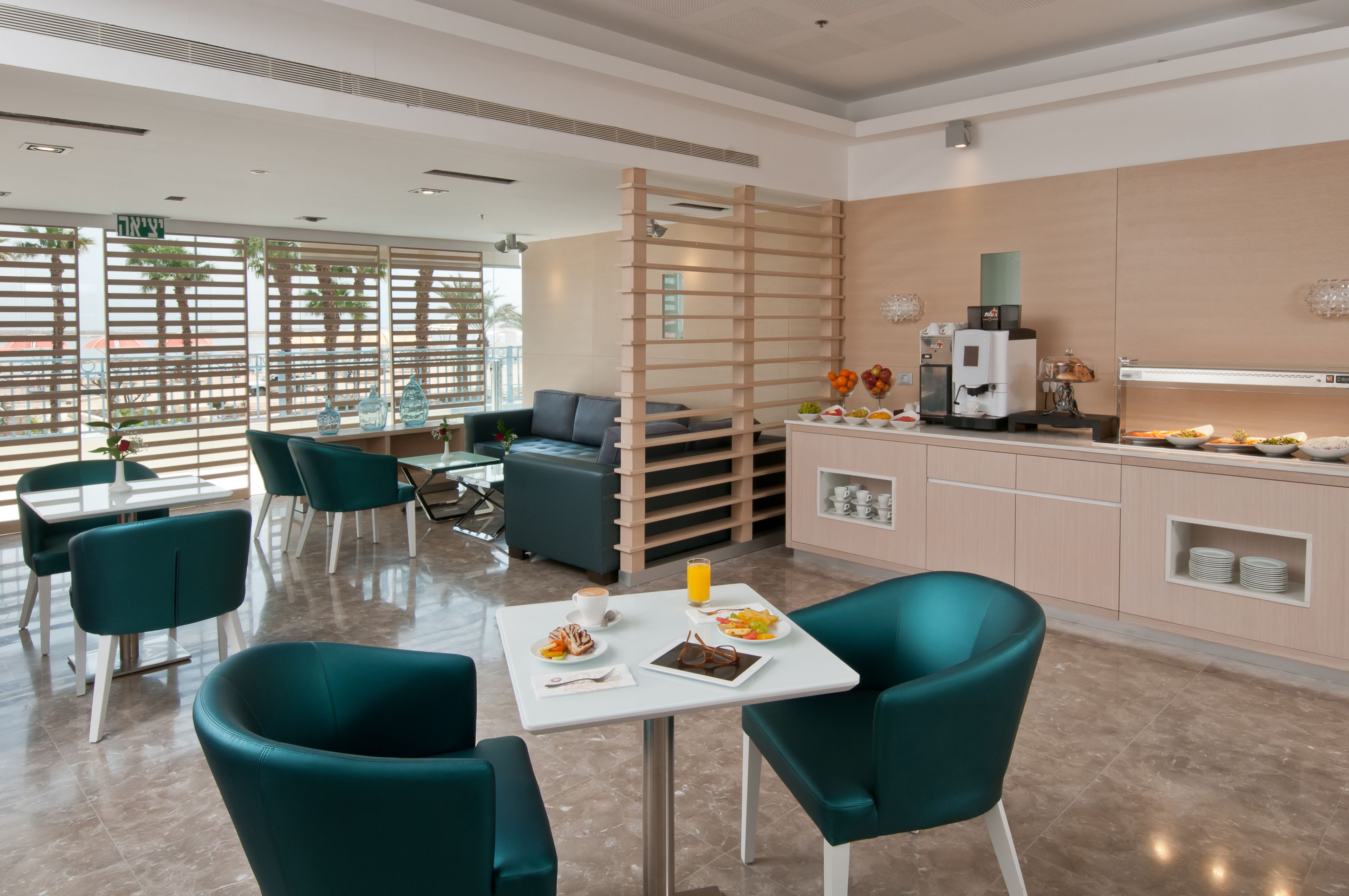 A modern dining area with teal chairs, a white table, and a kitchen area with a coffee machine and fruit display.
