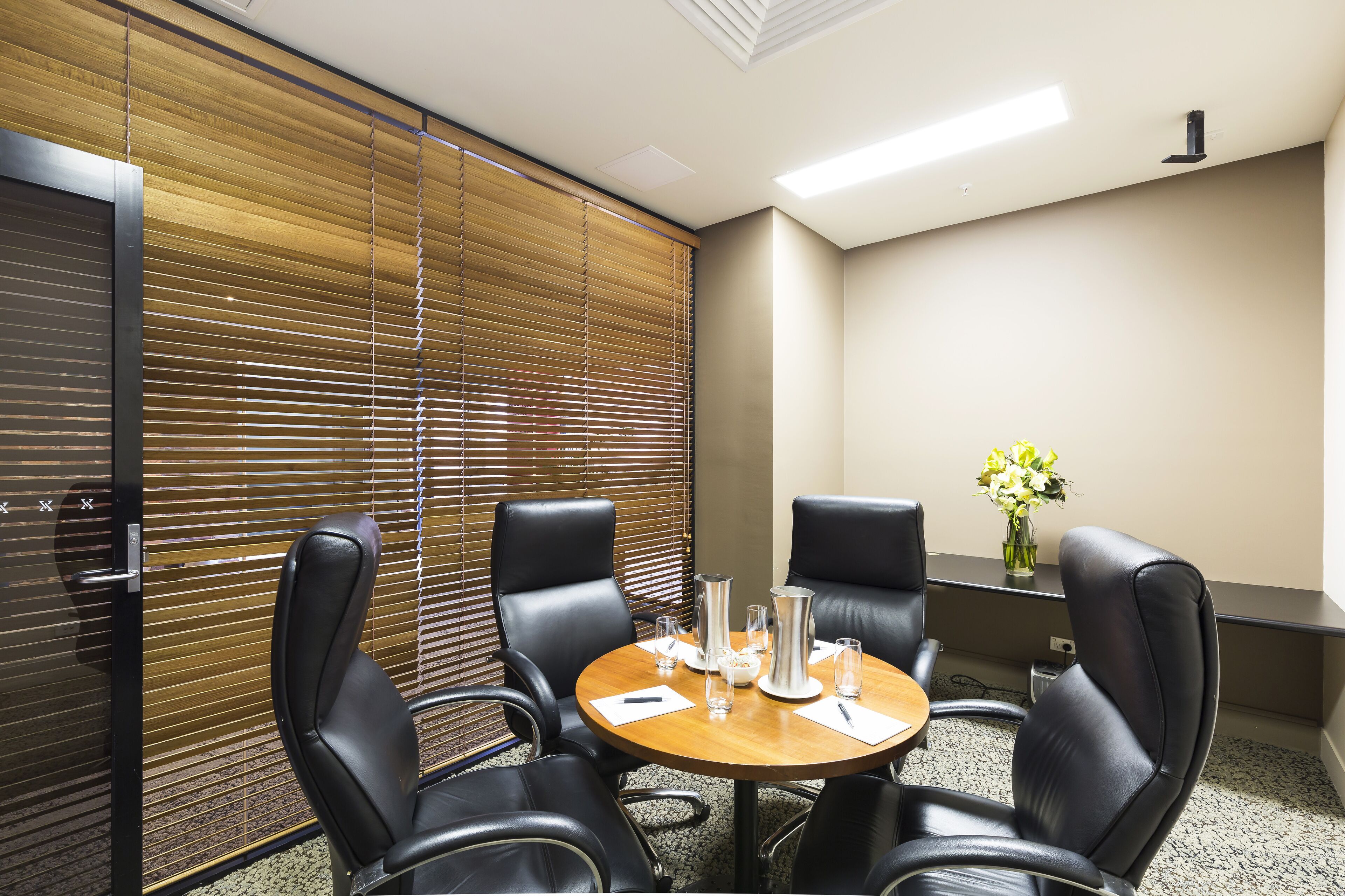 A conference room with a wooden table, four black leather chairs, a vase with flowers, and a window with blinds.