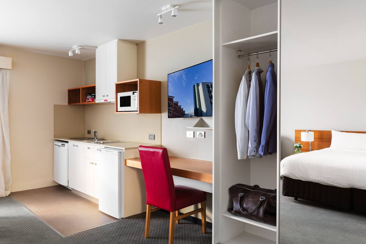 A compact hotel room with a kitchenette, a bed, a desk, and a red chair.