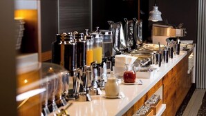 Una barra de desayuno con varias máquinas de café, un dispensador de jugo y una jarra de salsa roja.