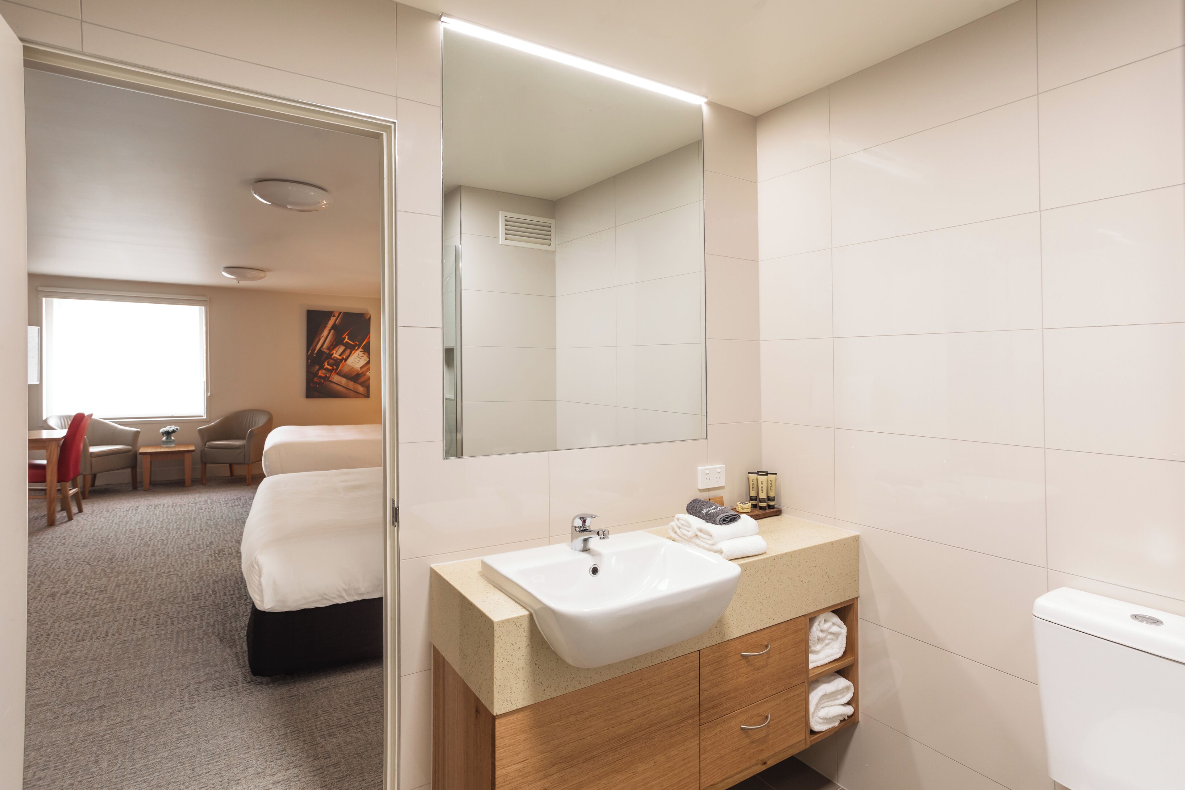 A hotel room with a bed, a bathroom with a sink and mirror, a small table with chairs, and a window.