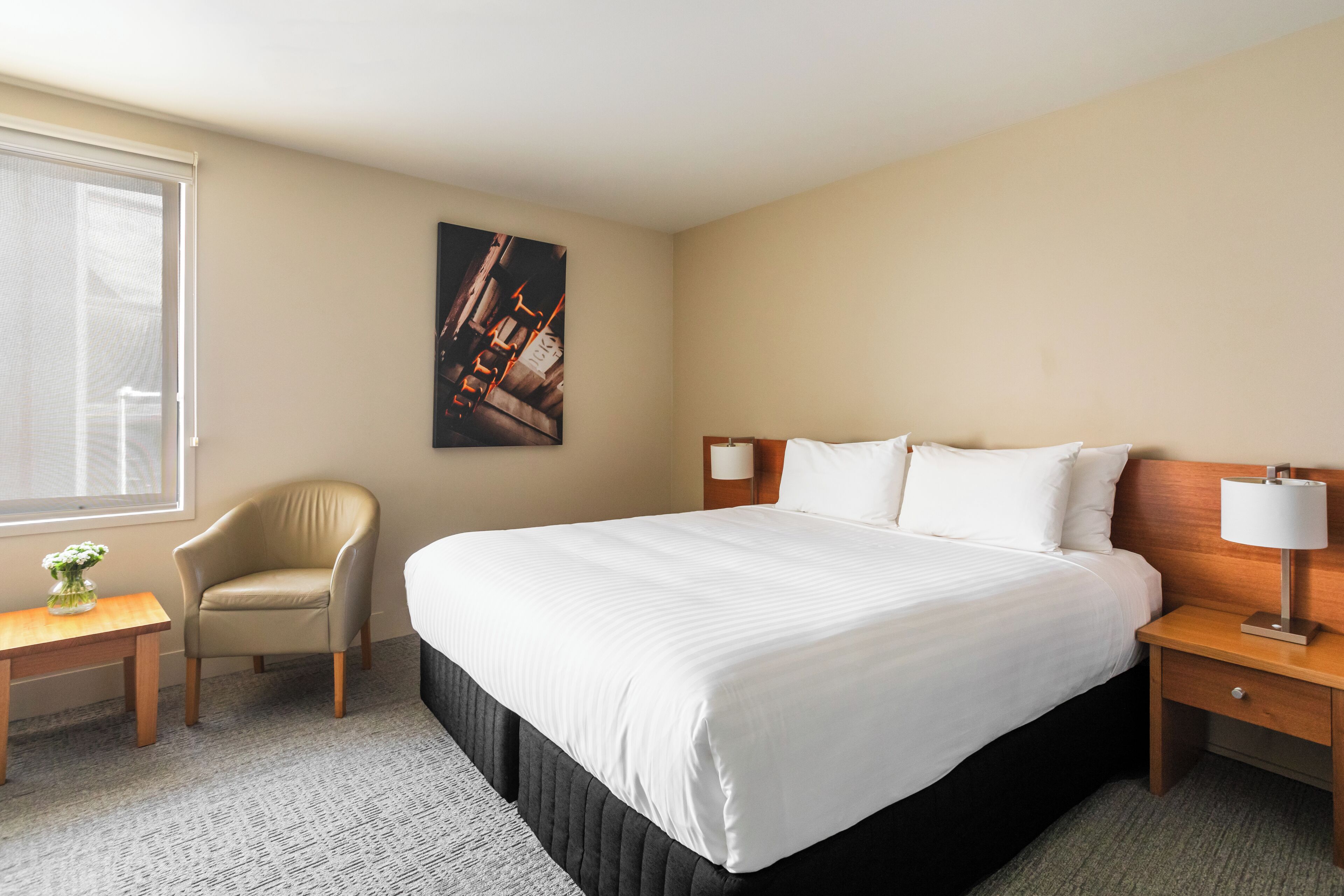 A hotel room with a large bed, a chair, a nightstand, a lamp, a picture on the wall, and a window.