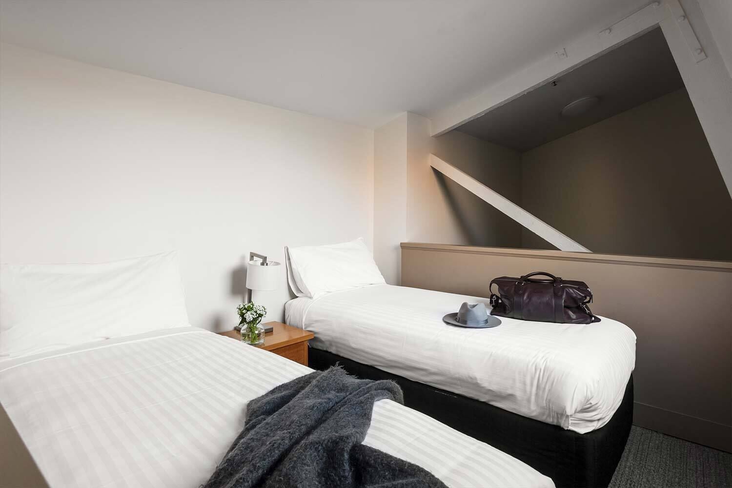 A hotel room with two beds, a nightstand with a lamp, a handbag, and a view of a staircase.