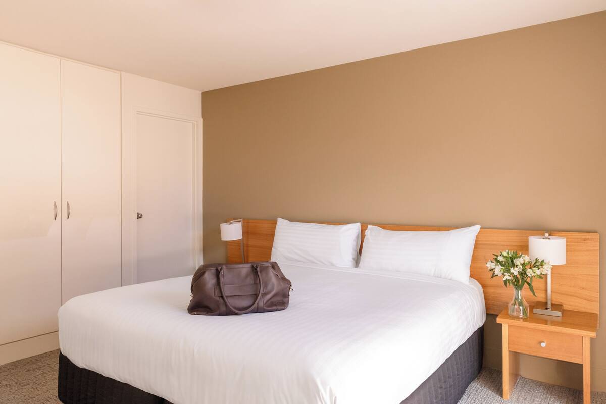 A hotel room with a bed, a nightstand with a lamp, a wooden headboard, and a wardrobe.