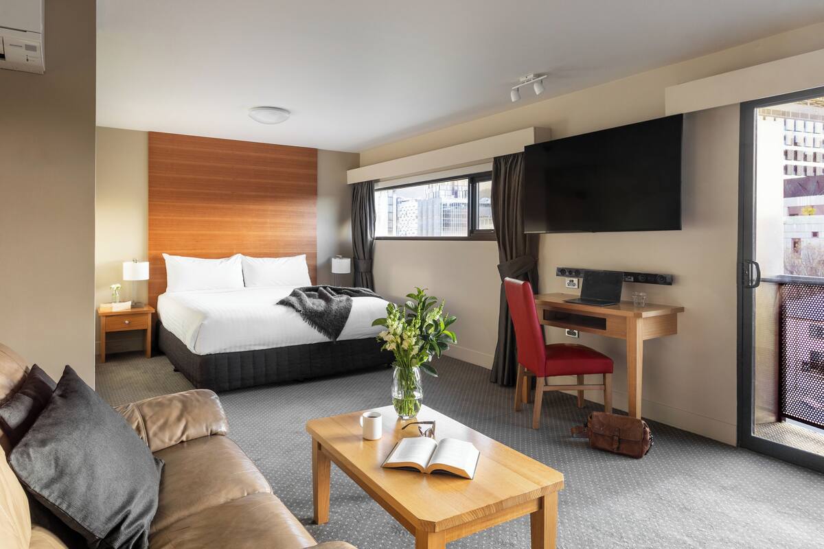 A hotel room with a bed, a desk with a laptop, a TV, a sofa, and a coffee table with a book and a vase.