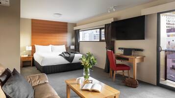 A hotel room with a bed, a desk with a laptop, a TV, a sofa, and a coffee table with a book and a vase.