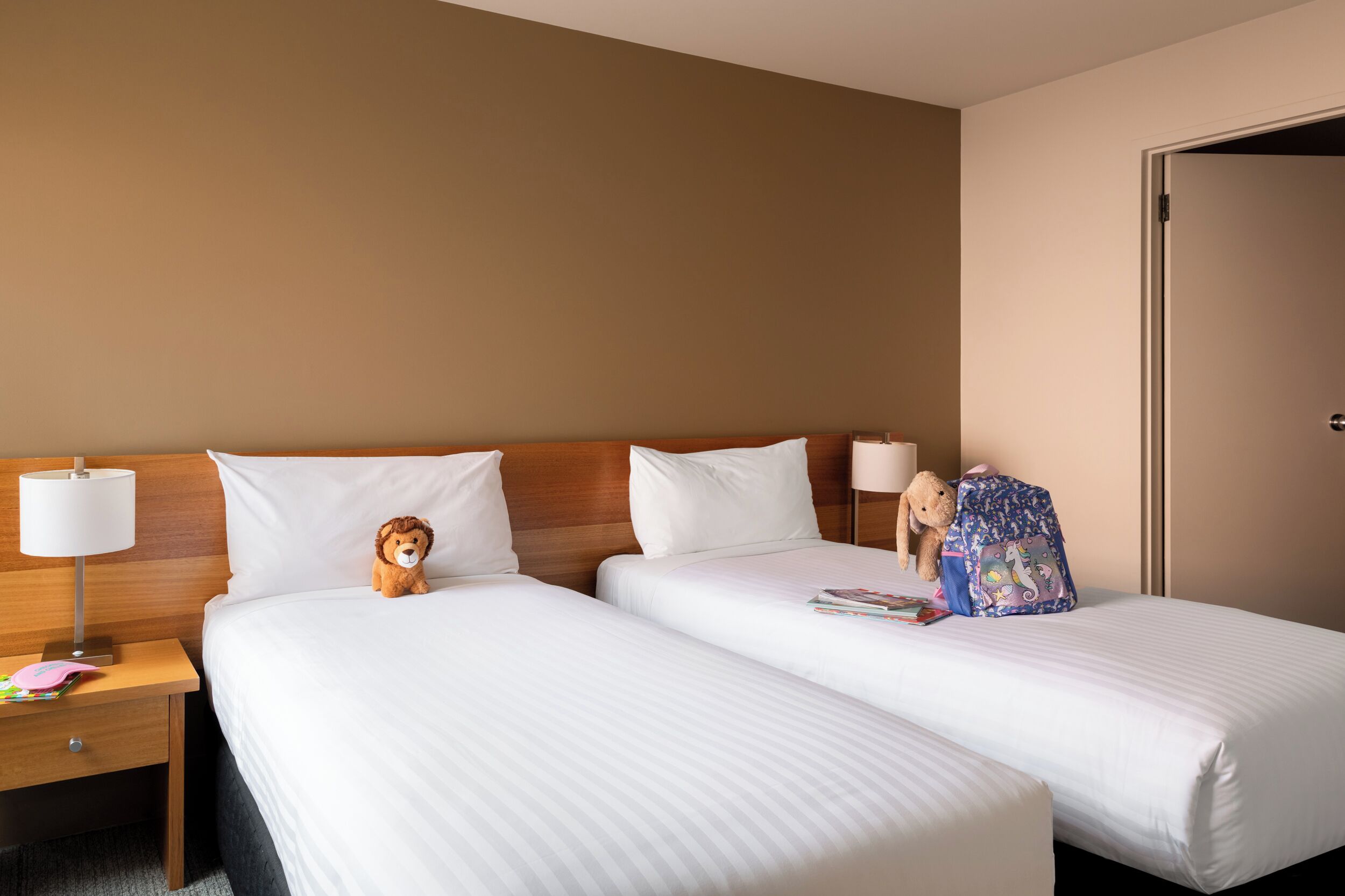 Ein Hotelzimmer mit zwei Einzelbetten, einem Holzkopfteil, einem Nachttisch mit Lampe und einem Teddybären auf einem Bett.