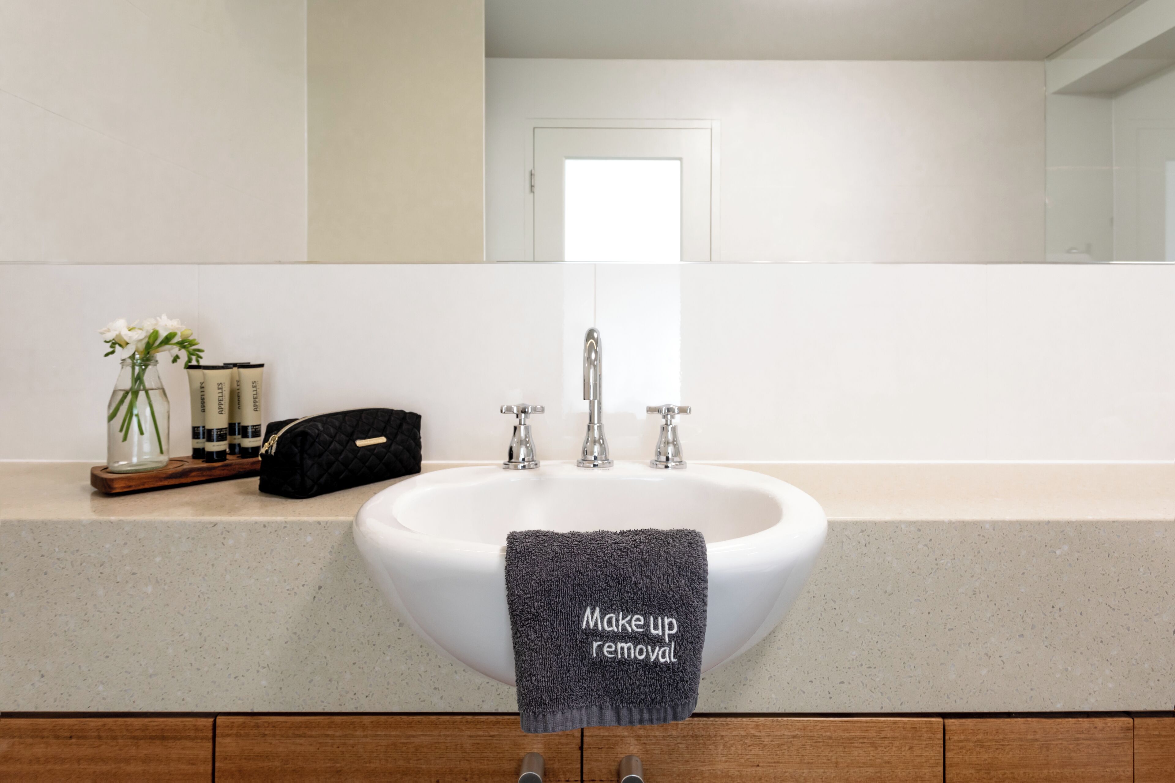 Un baño con un lavabo, una toalla con la etiqueta 'eliminador de maquillaje', un florero con flores y un espejo.