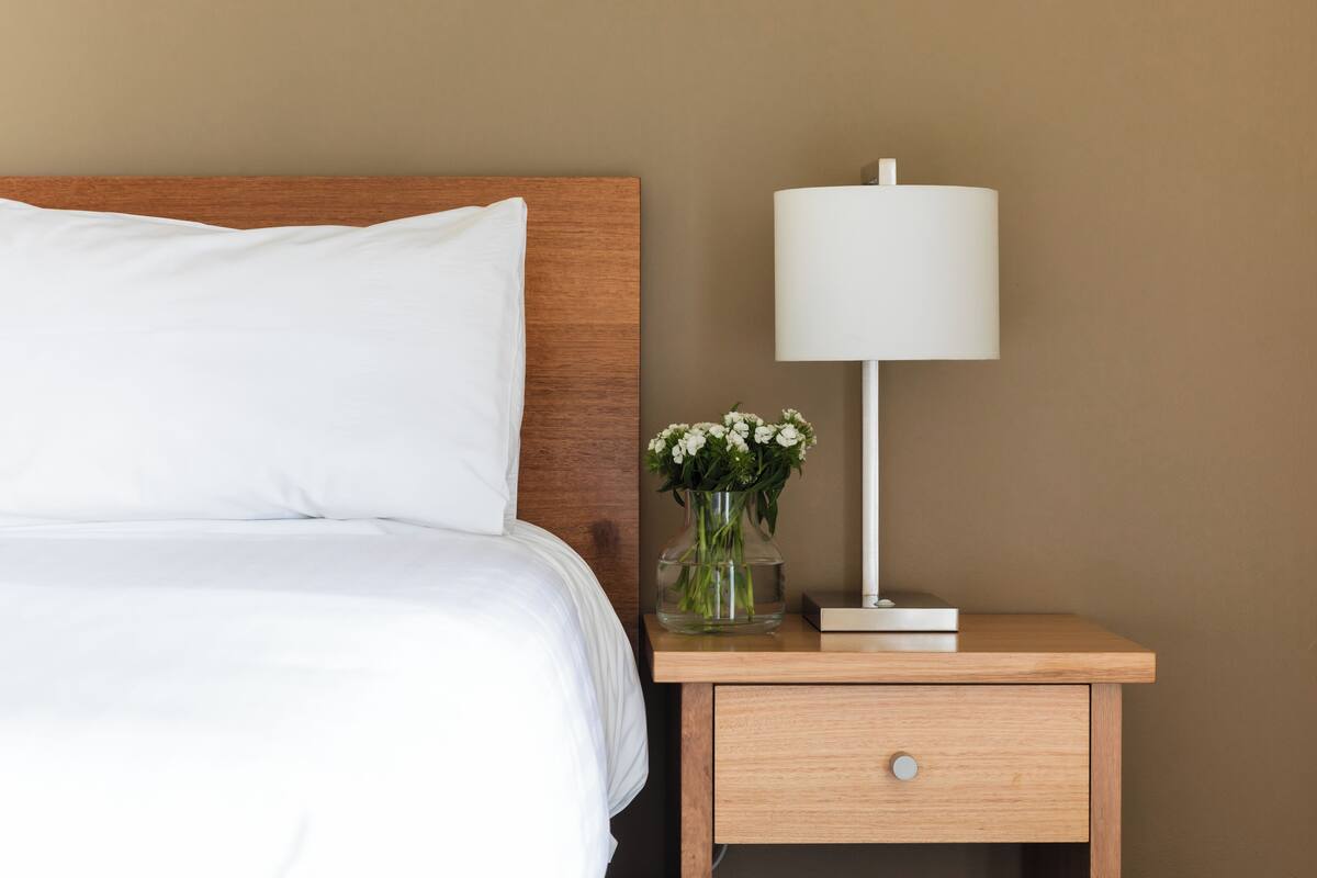 A neatly made bed with white linens, a wooden headboard, a bedside lamp, and a vase with flowers on a wooden nightstand.