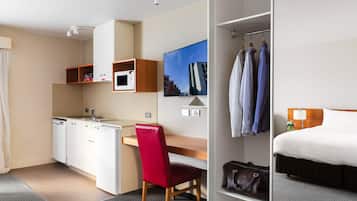 A compact hotel room with a kitchenette, a red chair, a bed, and a wardrobe with hanging clothes.